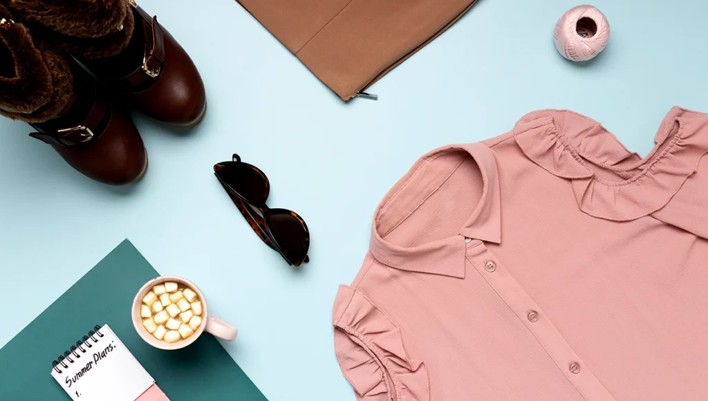 Flat lay photography showing a pink shirt, brown shoes, a coffee cup, and a notebook arranged artistically.